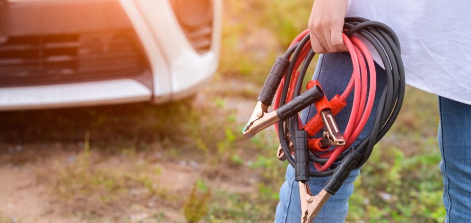 A woman holding jumper cables in front of a white car in a field of grass